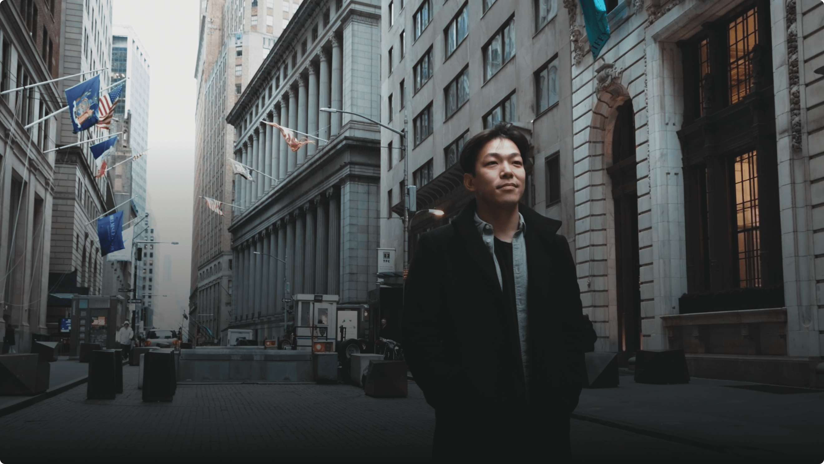 A person in a dark coat stands on a city street lined with tall buildings and flags, with hands in pockets, looking into the distance.